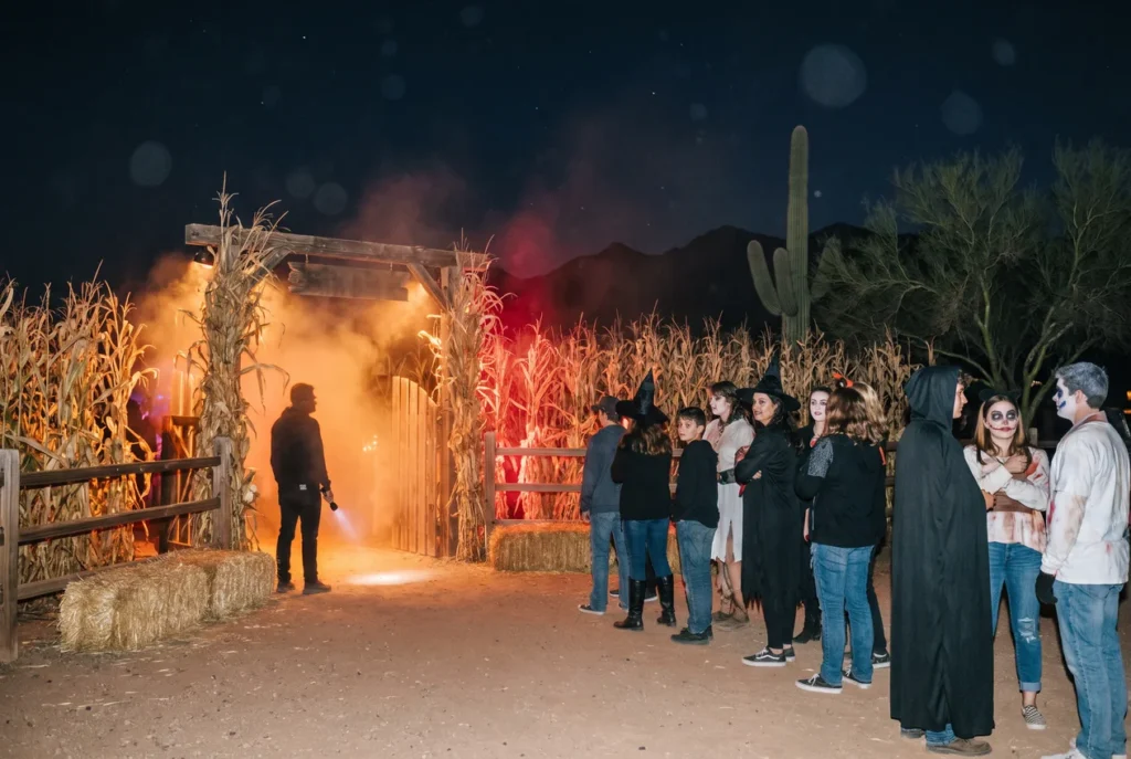 Costumed visitors lining up at night at the entrance to a haunted corn maze on a desert farm, fog drifting from the tall corn under red and orange light